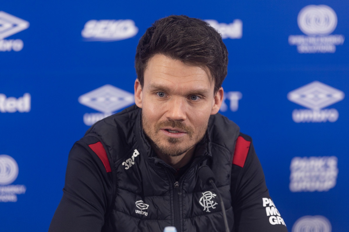 Danny Rohl sits at the desk in the Rangers press conference room at the training centre. Blue backdrop with sponsors logos on them. Danny wearing the black Rangers training gear.